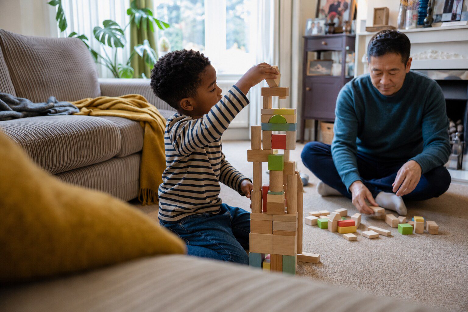 A foster carer sitting on the floor with a toddler, watching them stack coloured bricks.