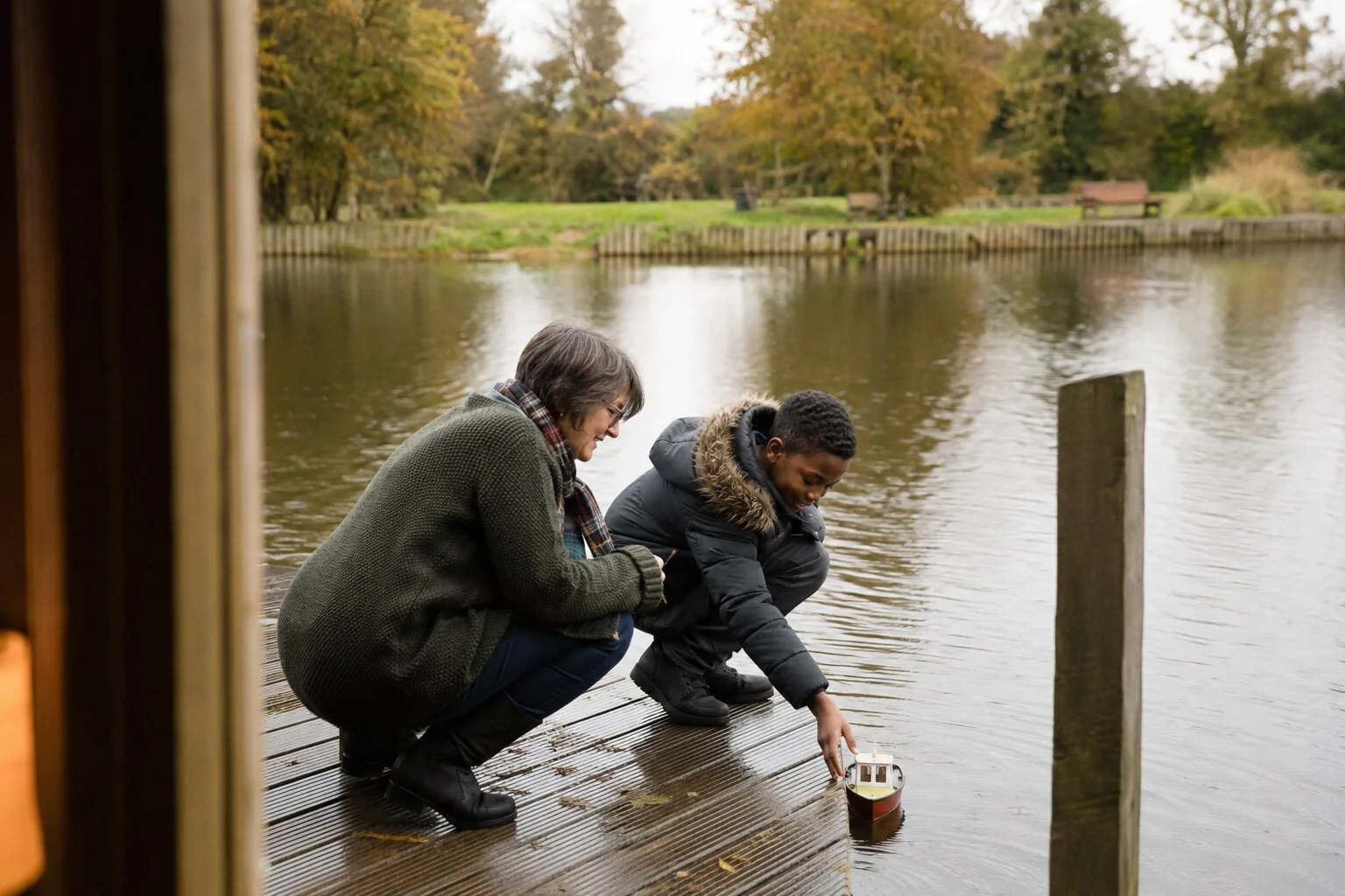 An adult and a young person trying a small toy boat at the edge of a pond