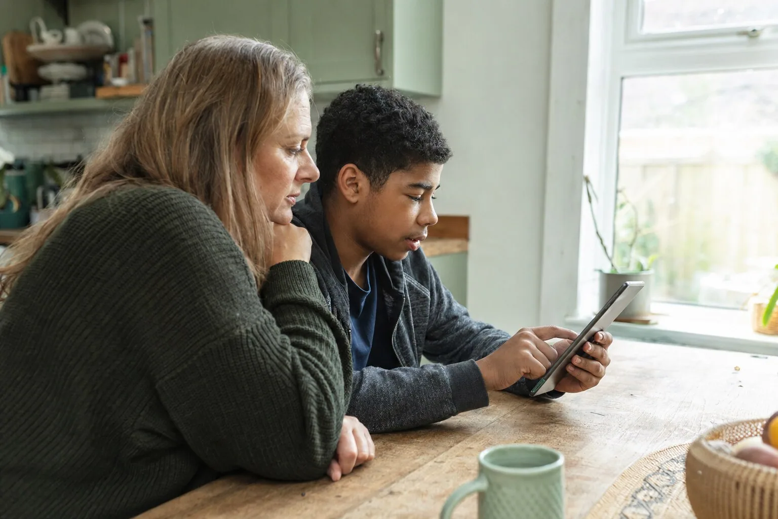 An adult and a teenager looking at a tablet together at the kitchen table