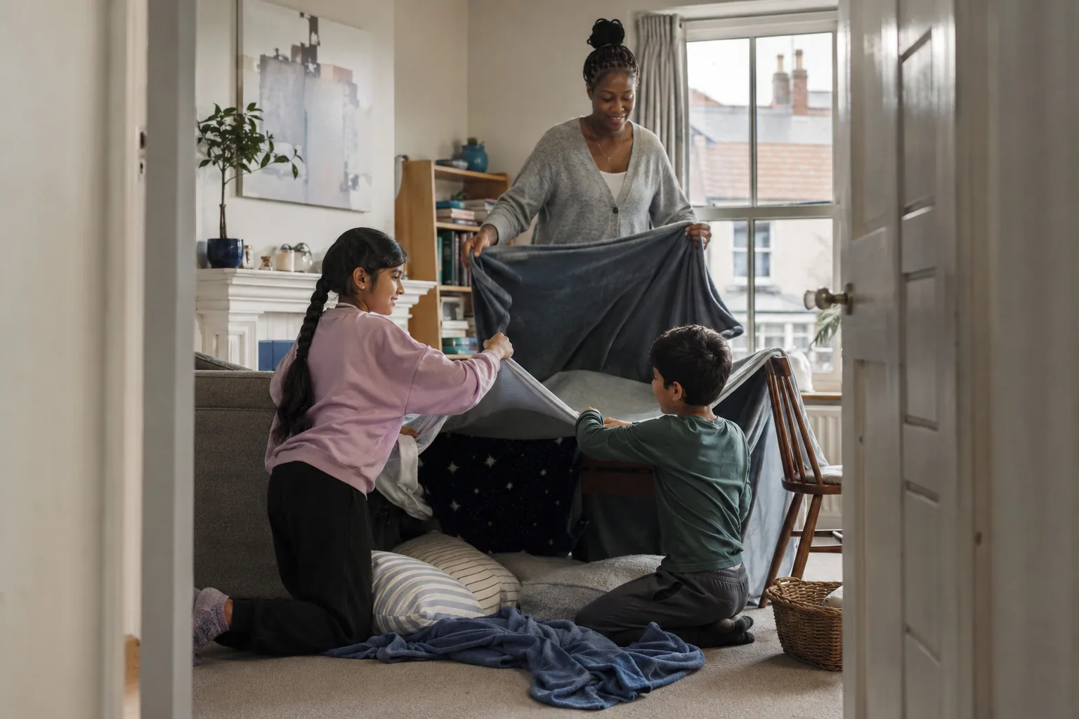 An adult helping two children make a blanket den in the living room