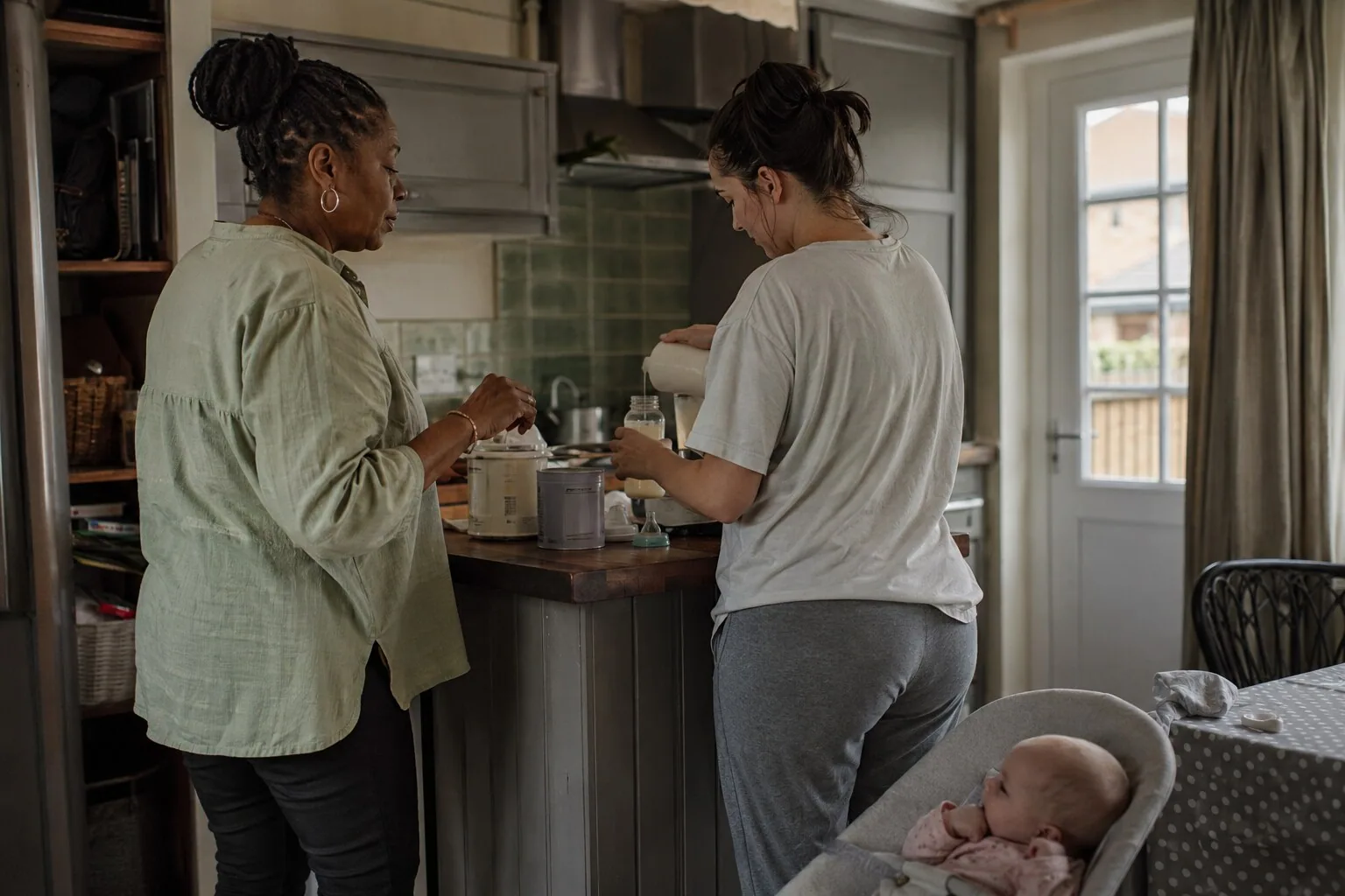 Two women preparing a drink in the kitchen while a baby rests nearby